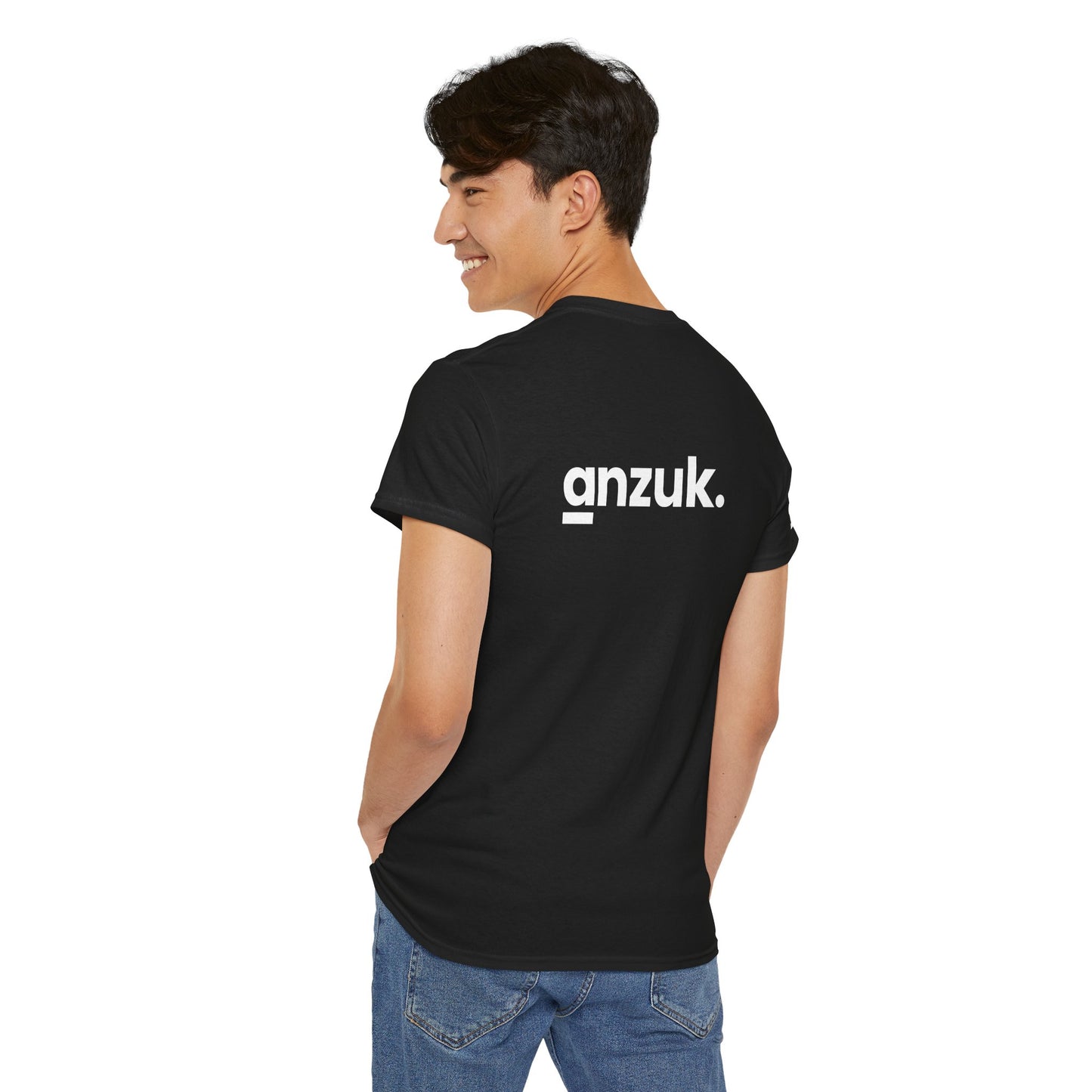 A person is standing and smiling, with their back partially turned toward the camera. They are wearing a black T-shirt with the word anzuk. printed on the back, alongside blue jeans.